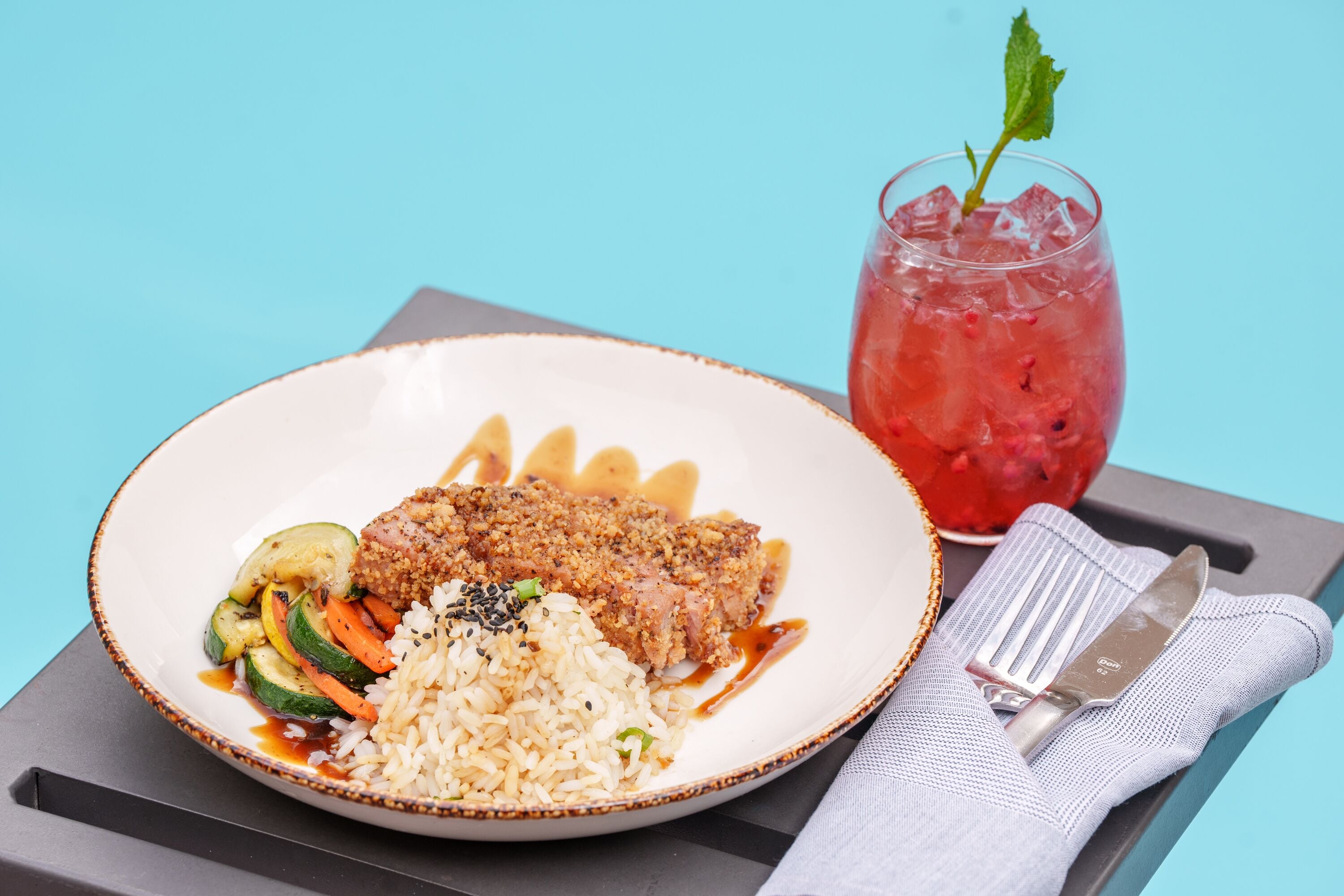 A plate of food with rice, vegetables, and a breaded item, accompanied by a drink with ice and a mint garnish.