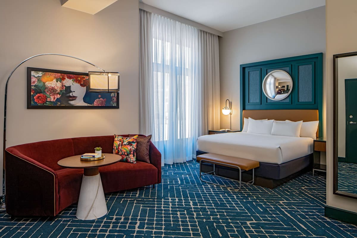 Habitación de hotel con sofá rojo, una mesita, cama con cabecera, un espejo grande y un cuadro en la pared.