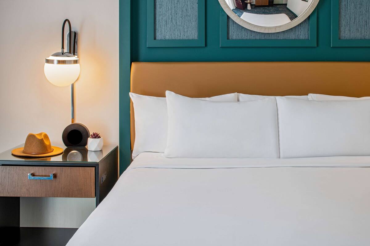Una cama bien hecha con sábanas blancas, una lámpara de noche, un sombrero y una pequeña planta en maceta sobre una mesita de madera.