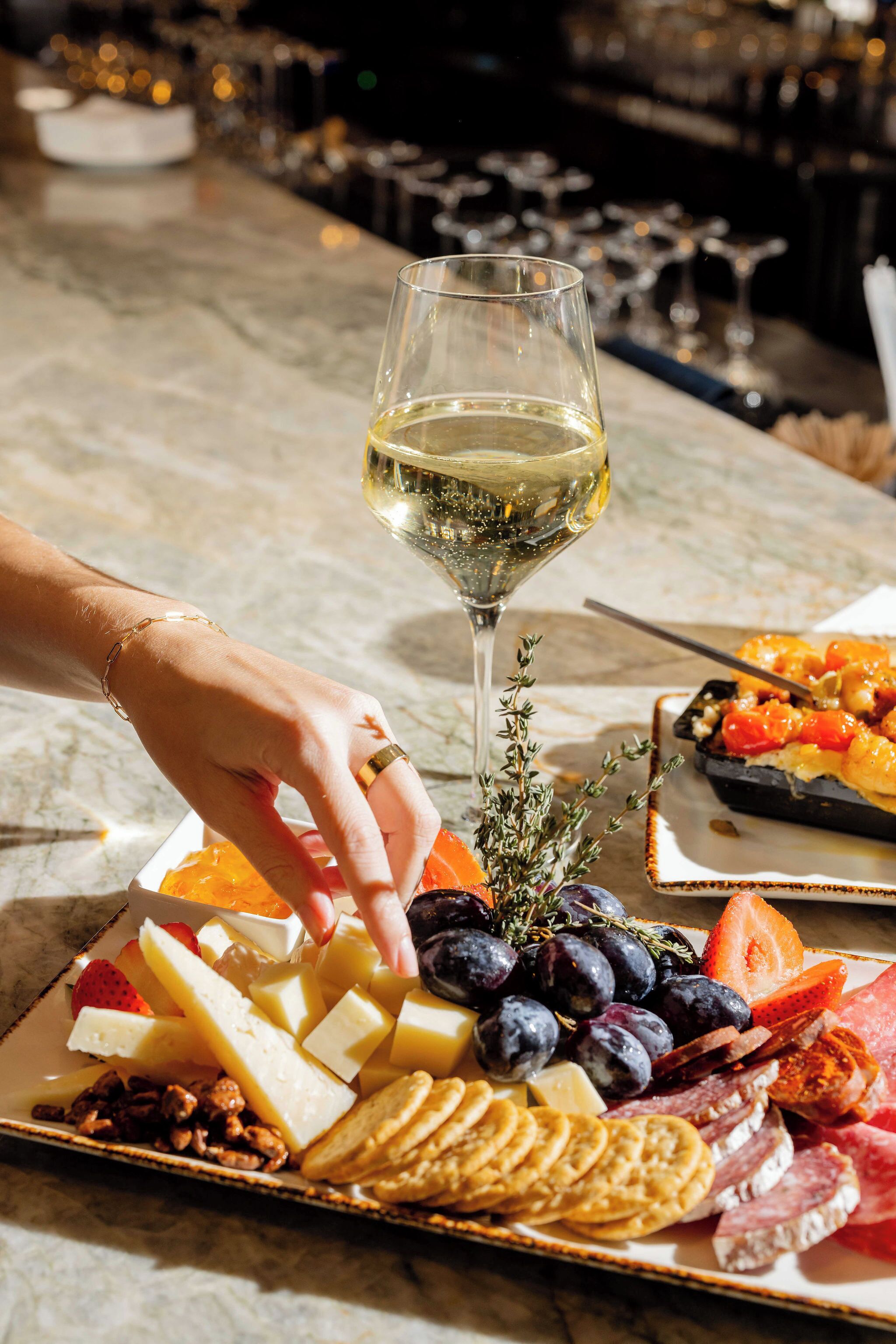 A charcuterie board with cheese, fruits, and nuts, and a glass of white wine on a marble table.