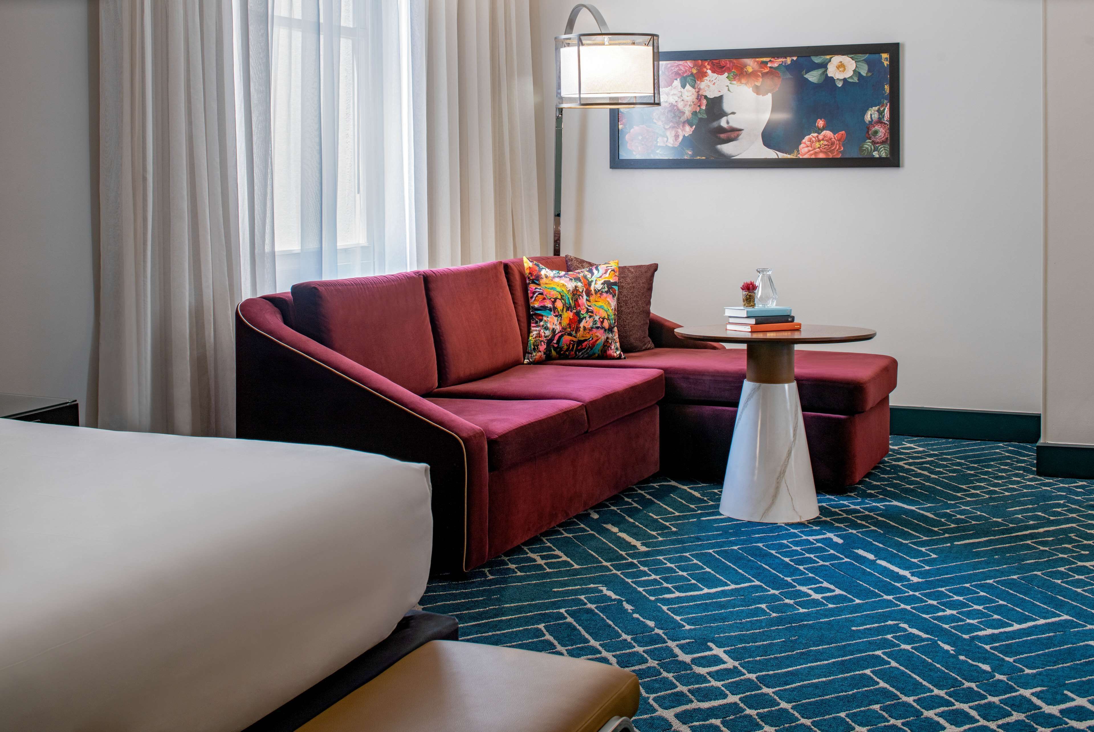 A hotel room with a red sofa, a small round table with books, a lamp, and a painting on the wall.