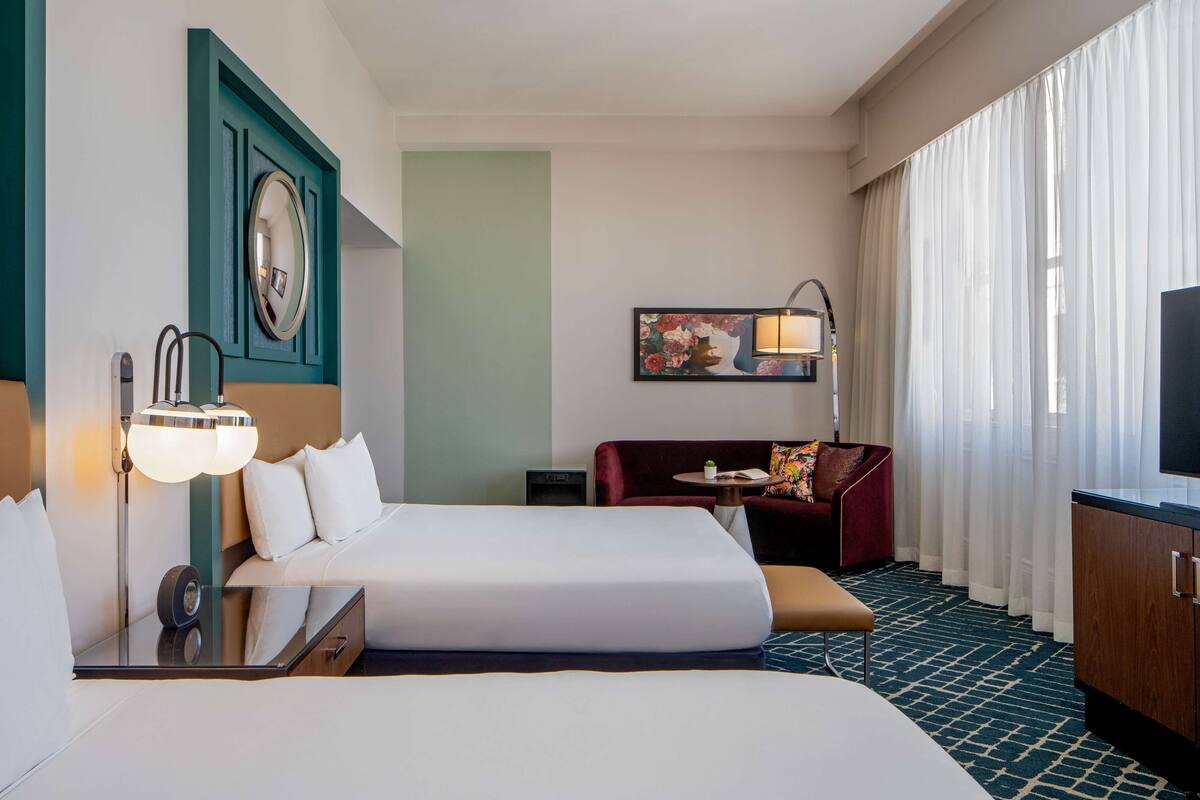 Habitación de hotel con una cama grande, un escritorio con lámpara, una mesita con silla, un sofá y un televisor.