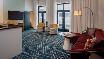 A hotel room with a blue patterned carpet, a red sofa, two armchairs, a small table with books, a TV, and a large window with curtains.
