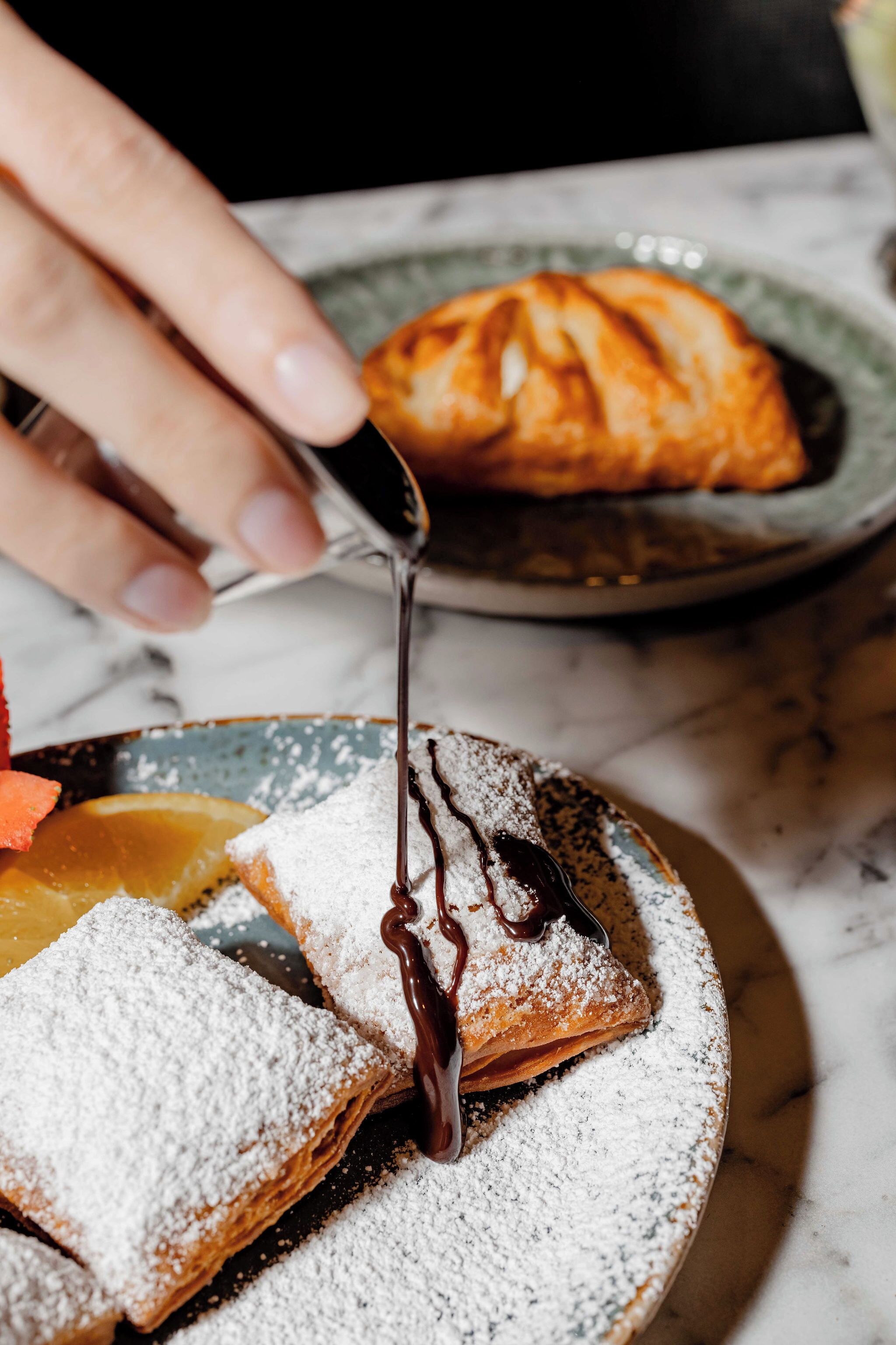 Ein Dessertteller mit Puderzucker und Schokoladensauce, im Hintergrund ein Croissant.