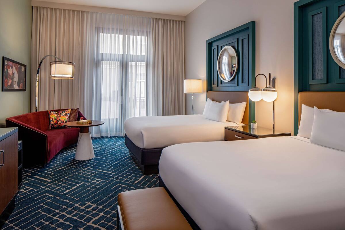 Habitación de hotel con dos camas, un sofá rojo, una mesita pequeña y un ventanal con cortinas traslúcidas.