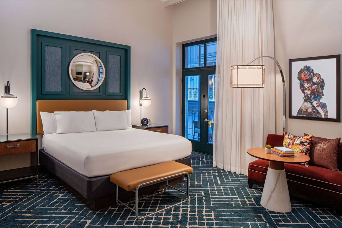 Habitación de hotel con una cama grande, un banco, una mesita, un sofá, un espejo, una lámpara y una ventana con vistas.