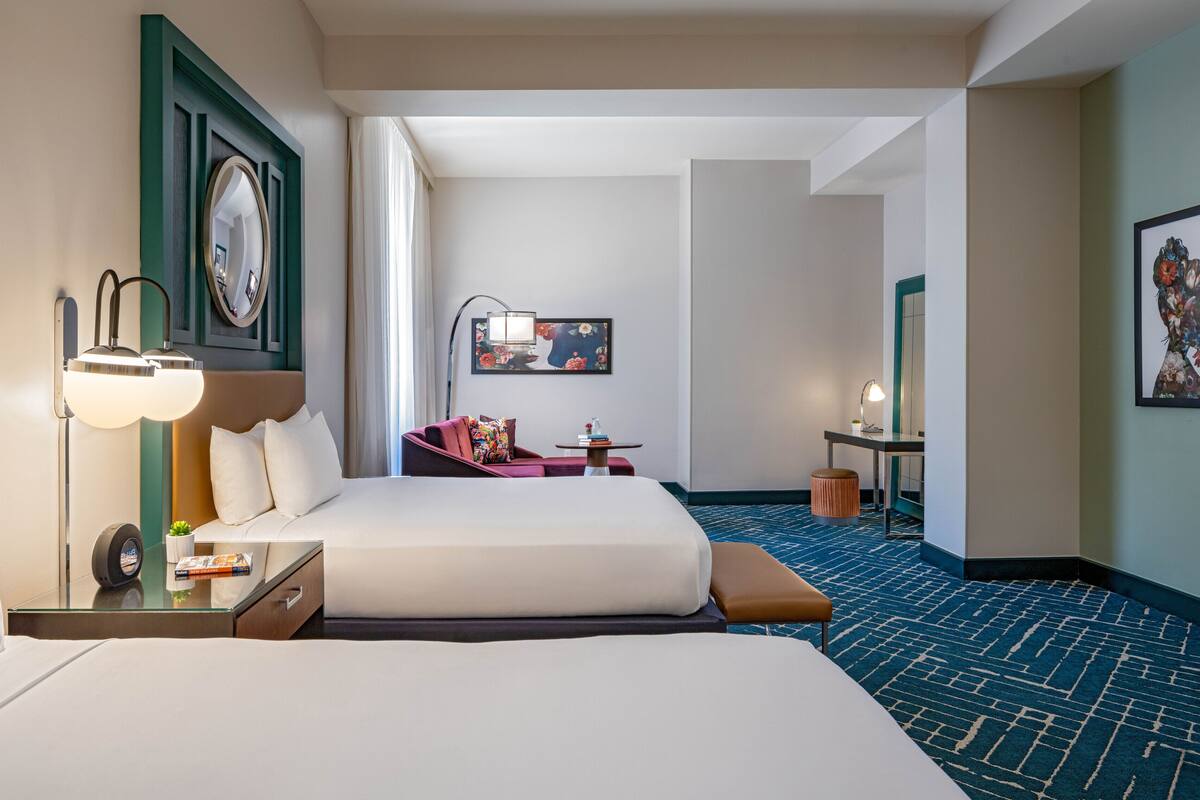 Habitación de hotel con dos camas, un escritorio, una silla, una lámpara y un cuadro en la pared.