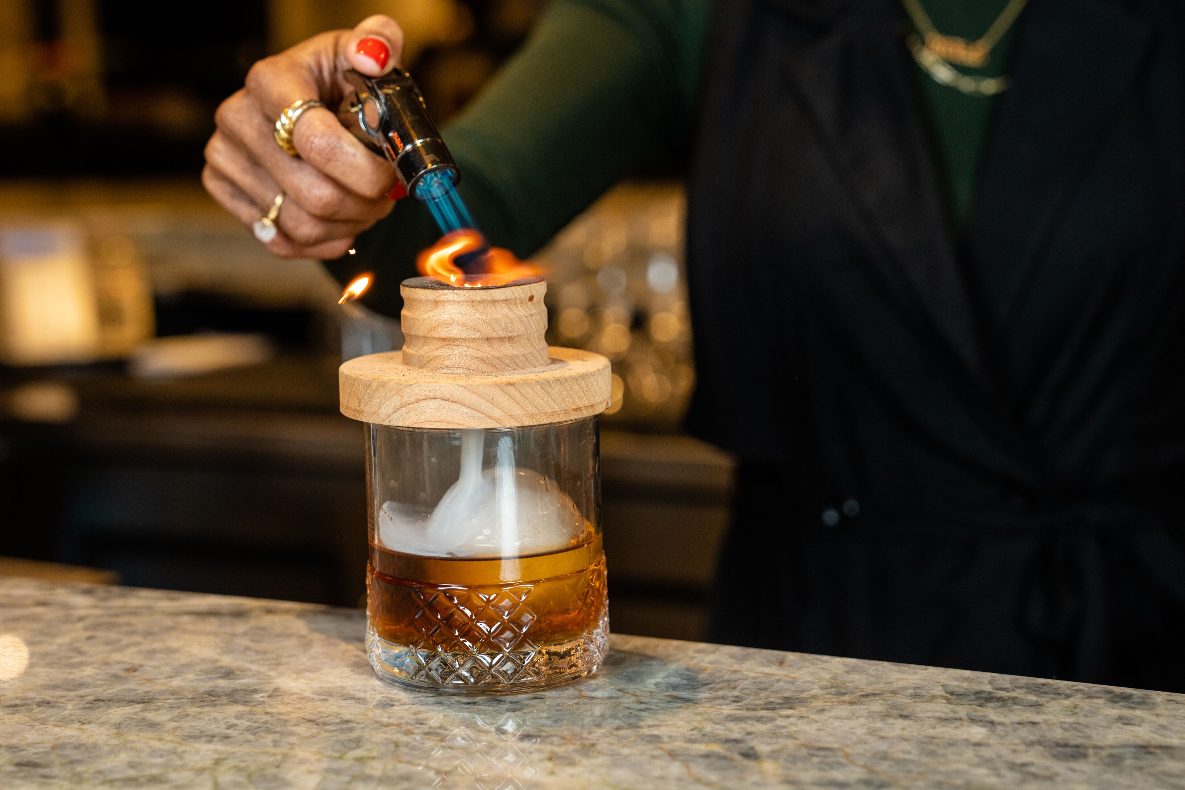 A hand holding a lighter with a flame, lighting a candle in a glass jar with a wooden lid.
