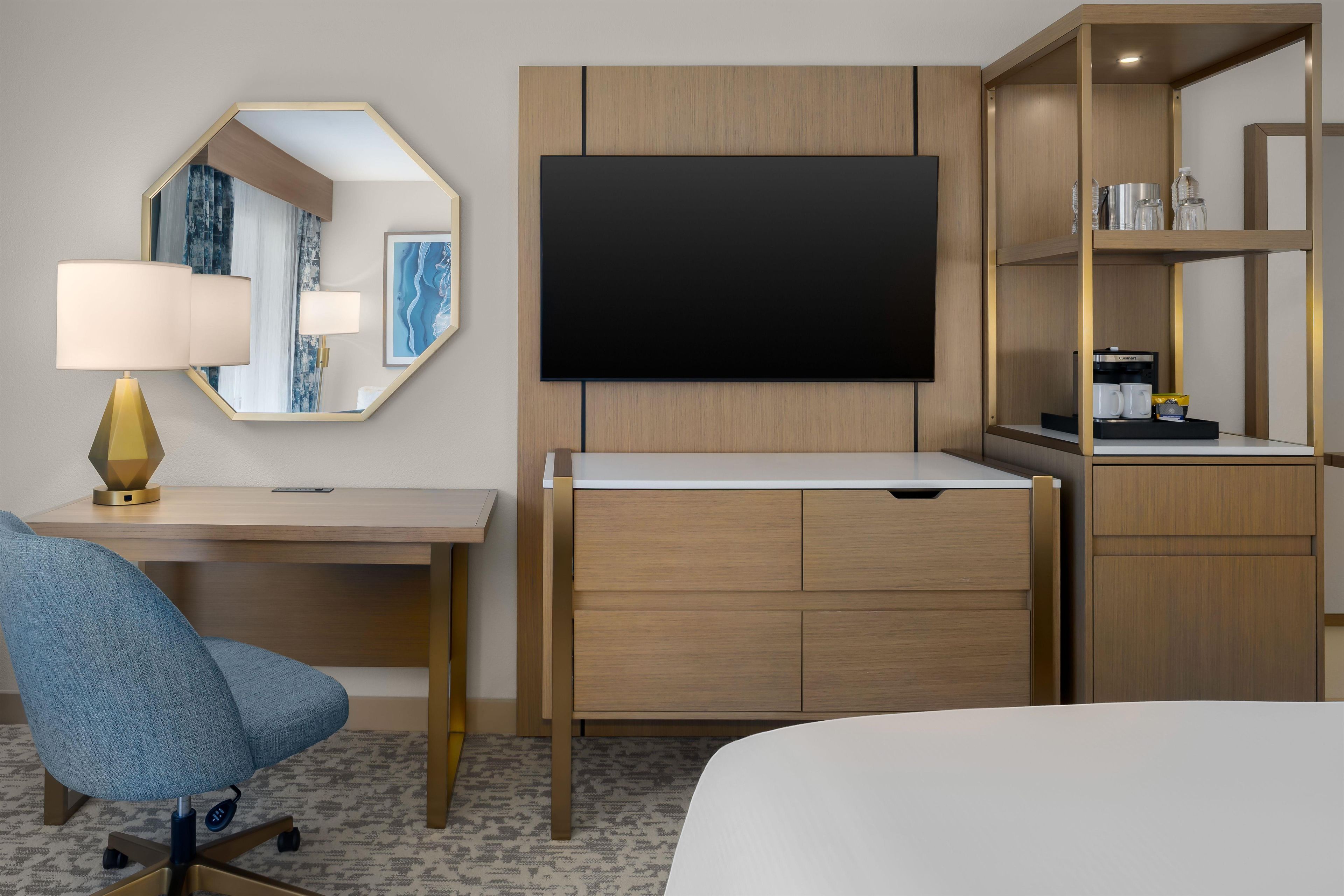 Habitación de hotel con escritorio, silla, cama, televisor y minibar incorporado.