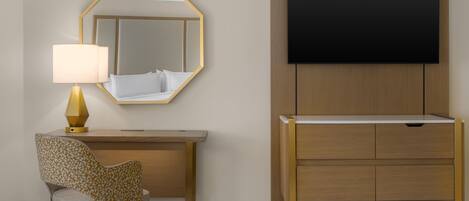 A hotel room with a bed, a desk with a lamp, a chair, a mirror, and a television.