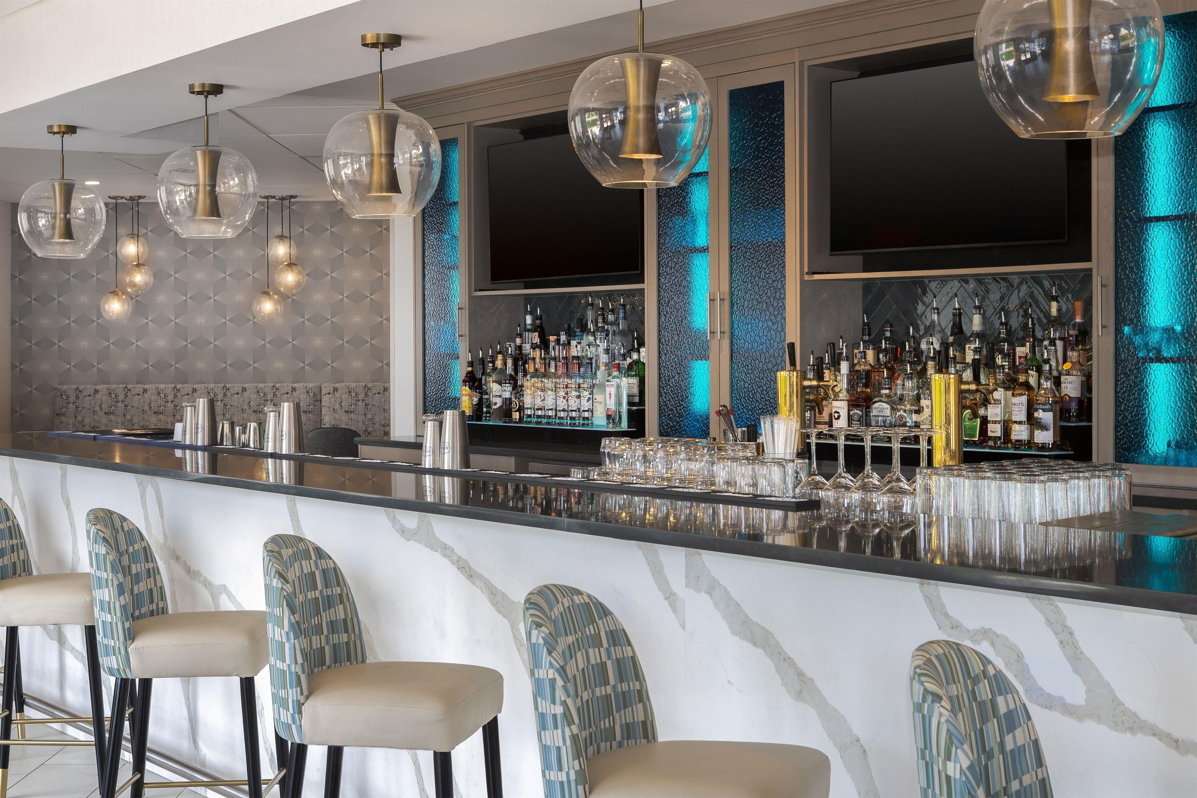 Zona de bar con mostrador de mármol, taburetes y una variedad de botellas de licores.
