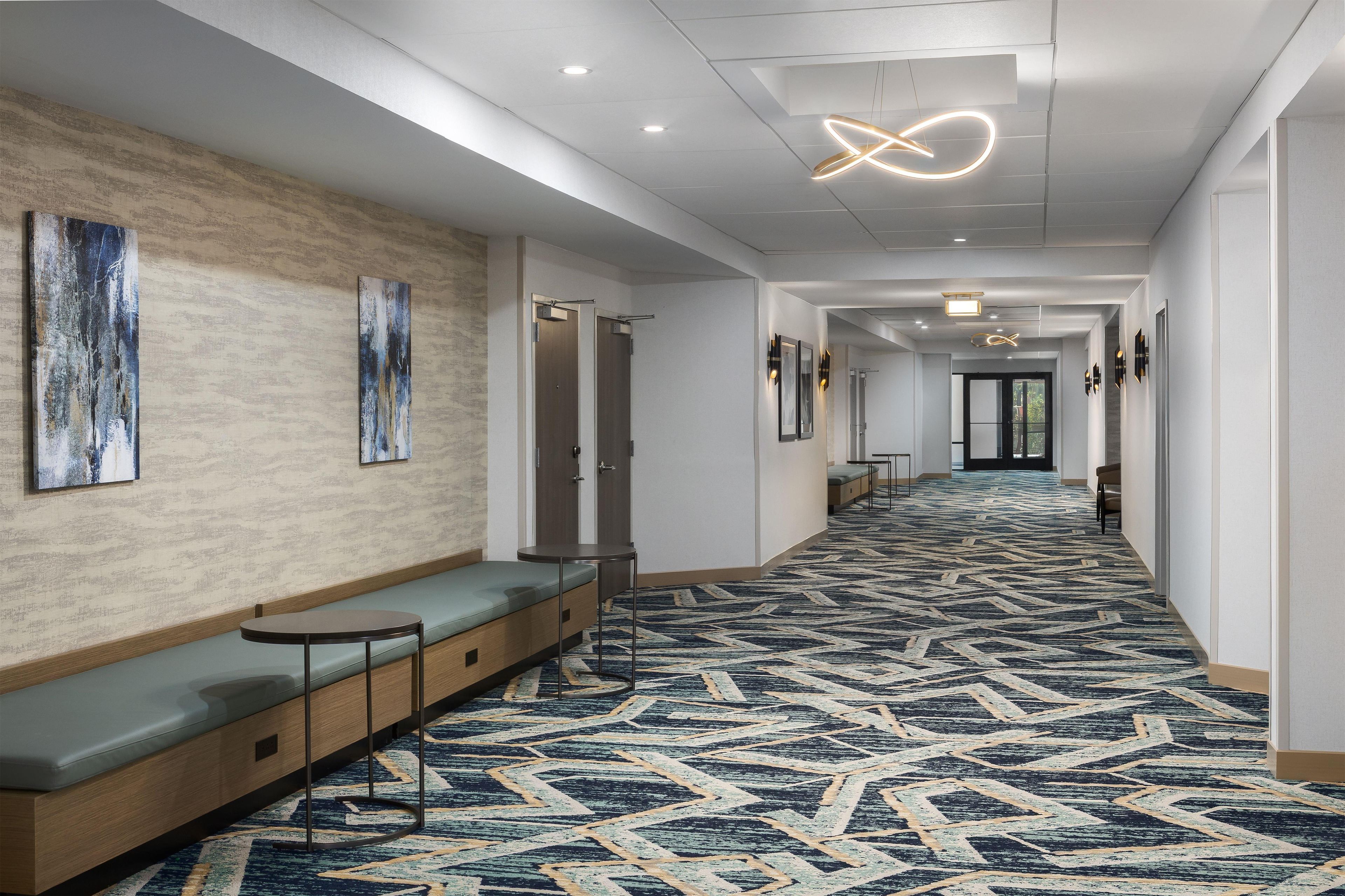 A hotel lobby with a patterned carpet, seating area, artwork, and a modern light fixture.