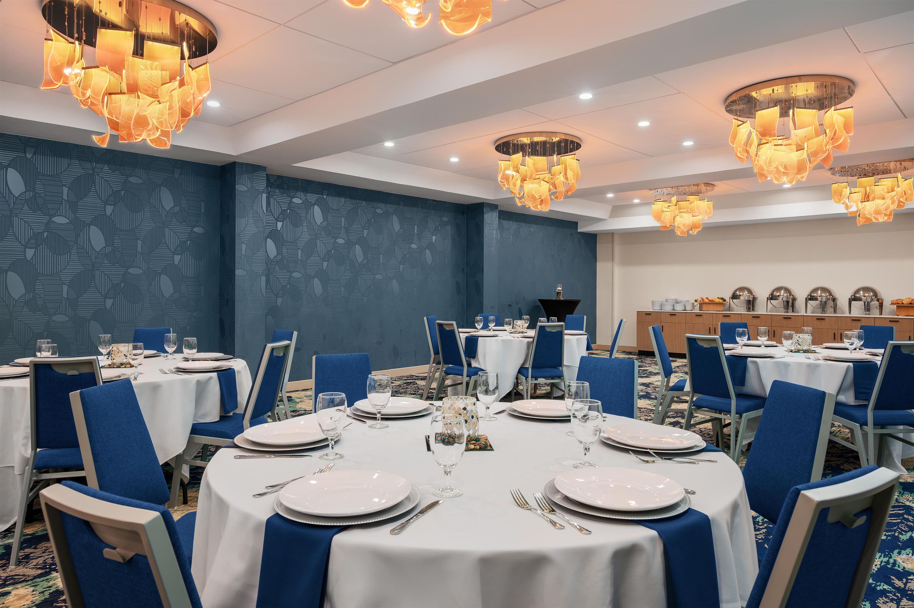 Ein Hotelrestaurant mit runden Tischen, blauen Stühlen und modernen Kronleuchtern.