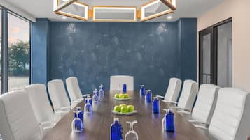 A conference room with a long table, white chairs, blue bottles, and a plate of green apples.