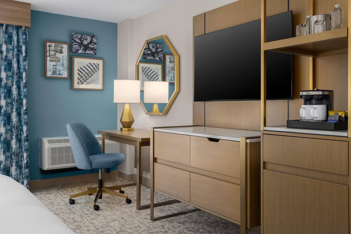 A hotel room with a desk, chair, flat-screen TV, coffee maker, and artwork on the wall.