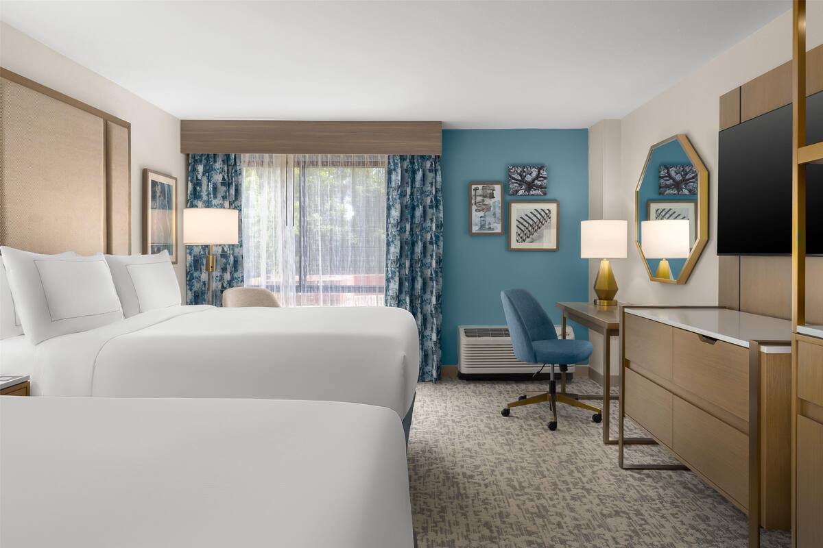 A hotel room with two beds, a desk, a chair, a TV, and a window with curtains.
