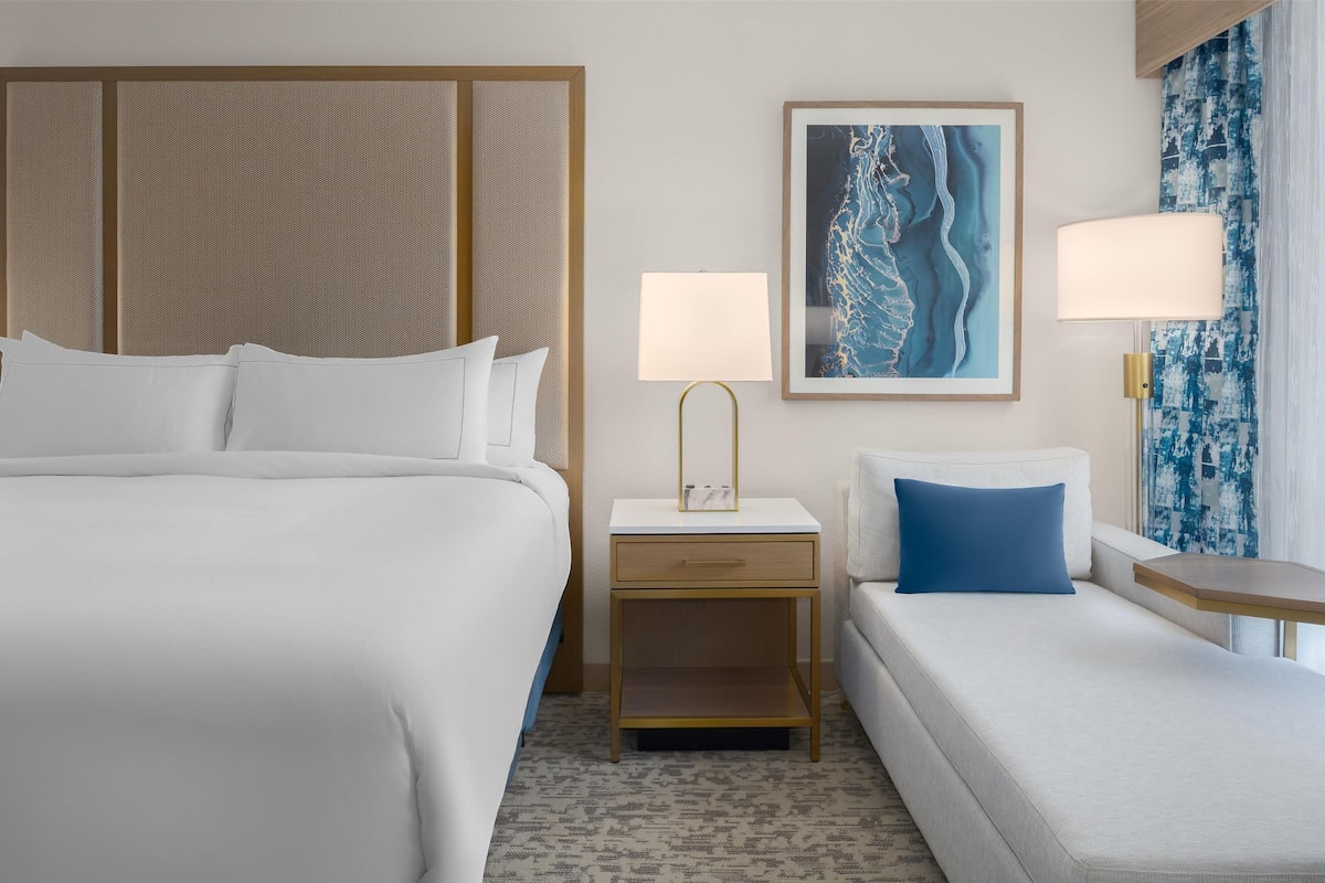 A hotel room with a large bed, a smaller sofa, a nightstand with a lamp, and a framed picture on the wall.