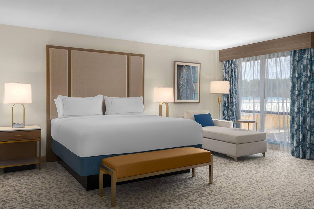 A hotel room with a large bed, bedside tables with lamps, a bench, a sofa, and a window with curtains.