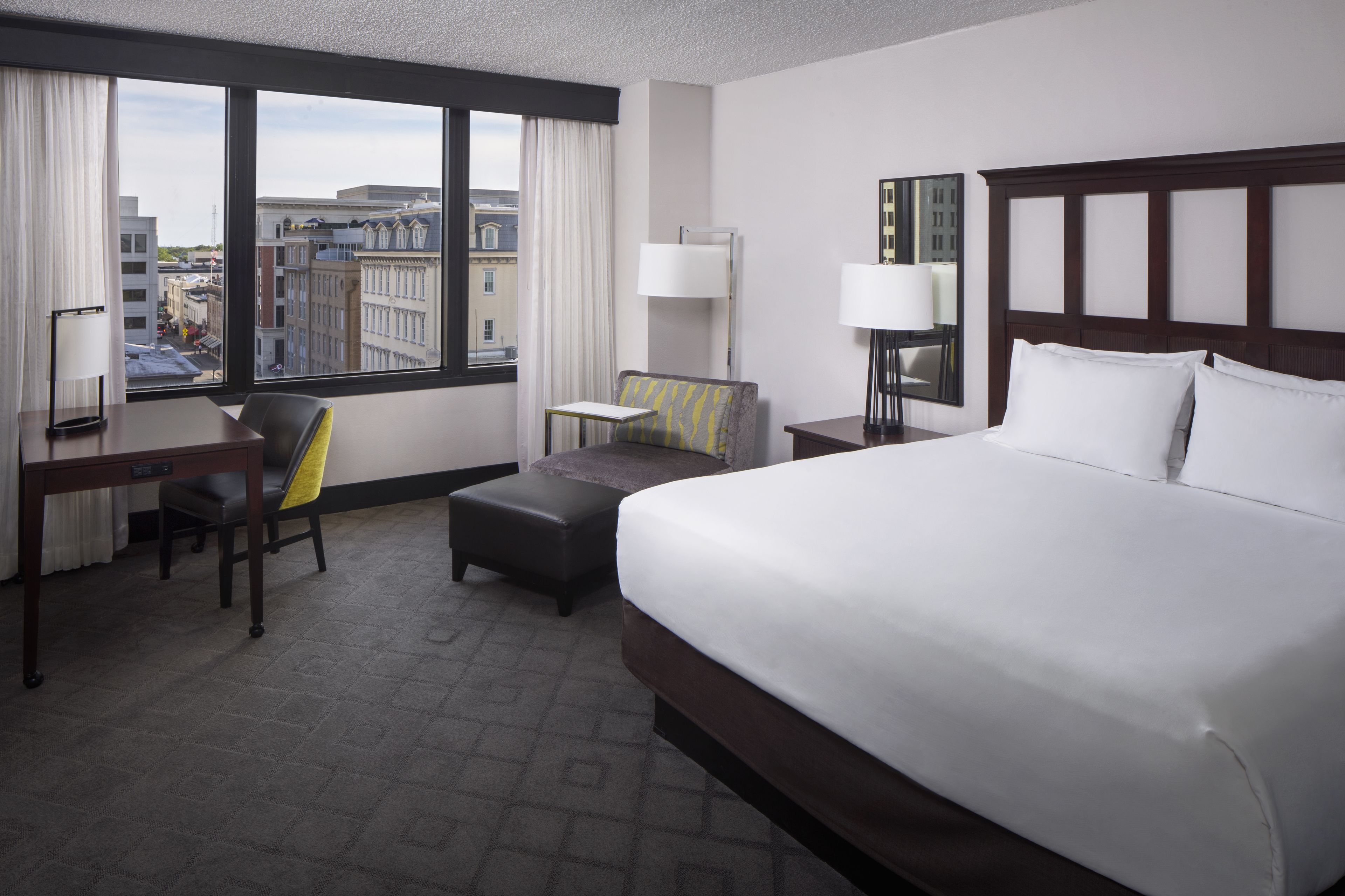 A hotel room with a large bed, a desk, a chair, a small table, and a view of the city through large windows.
