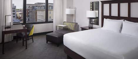 A hotel room with a large bed, a desk, a chair, a small table, and a view of the city through large windows.