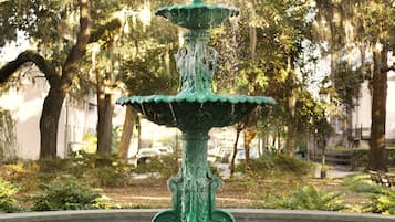 A multi-tiered green fountain with water cascading down, surrounded by a brick border and set in a park with trees and benches.