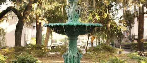 A multi-tiered green fountain with water cascading down, surrounded by a brick border and set in a park with trees and benches.
