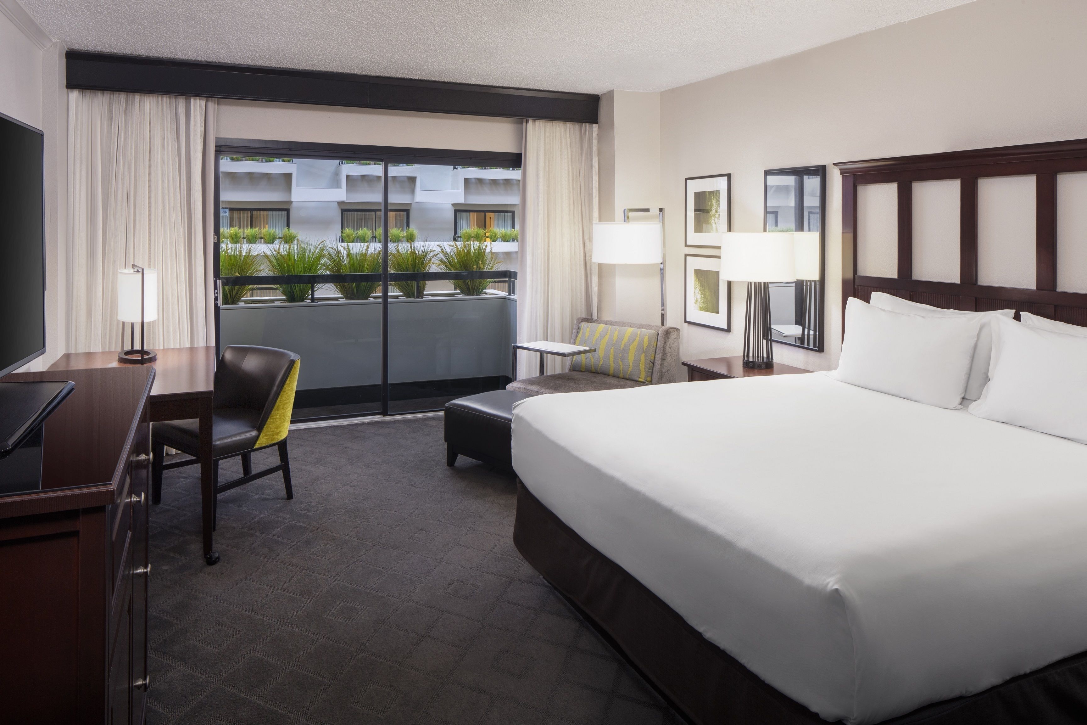 A hotel room with a large bed, a desk with a chair, a small table, and a sofa. There is a window with curtains and a view of a building with greenery outside.