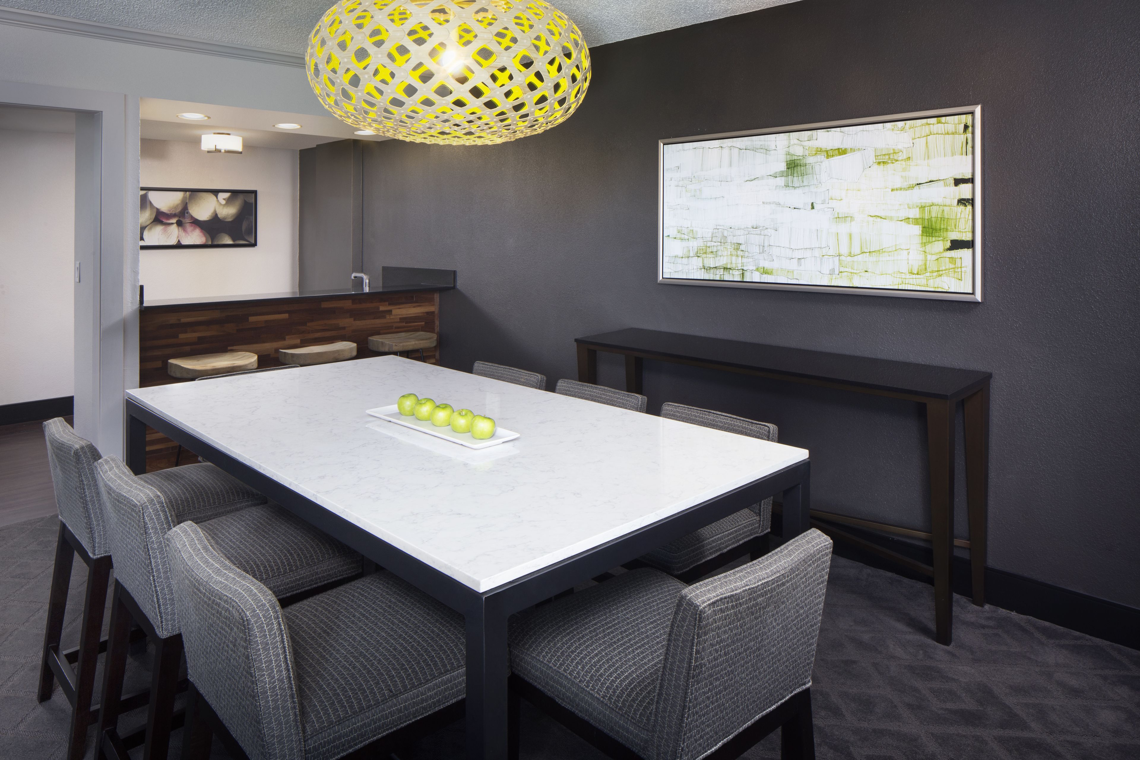 A modern dining area with a marble table, grey upholstered chairs, a yellow patterned pendant light, and a framed abstract painting on the wall.