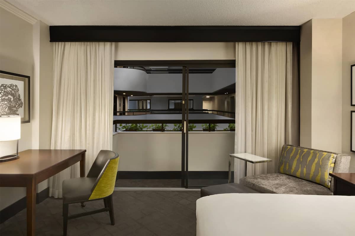 A hotel room with a bed, a desk, a chair, a sofa, and a view of a kitchen and dining area through a glass door.