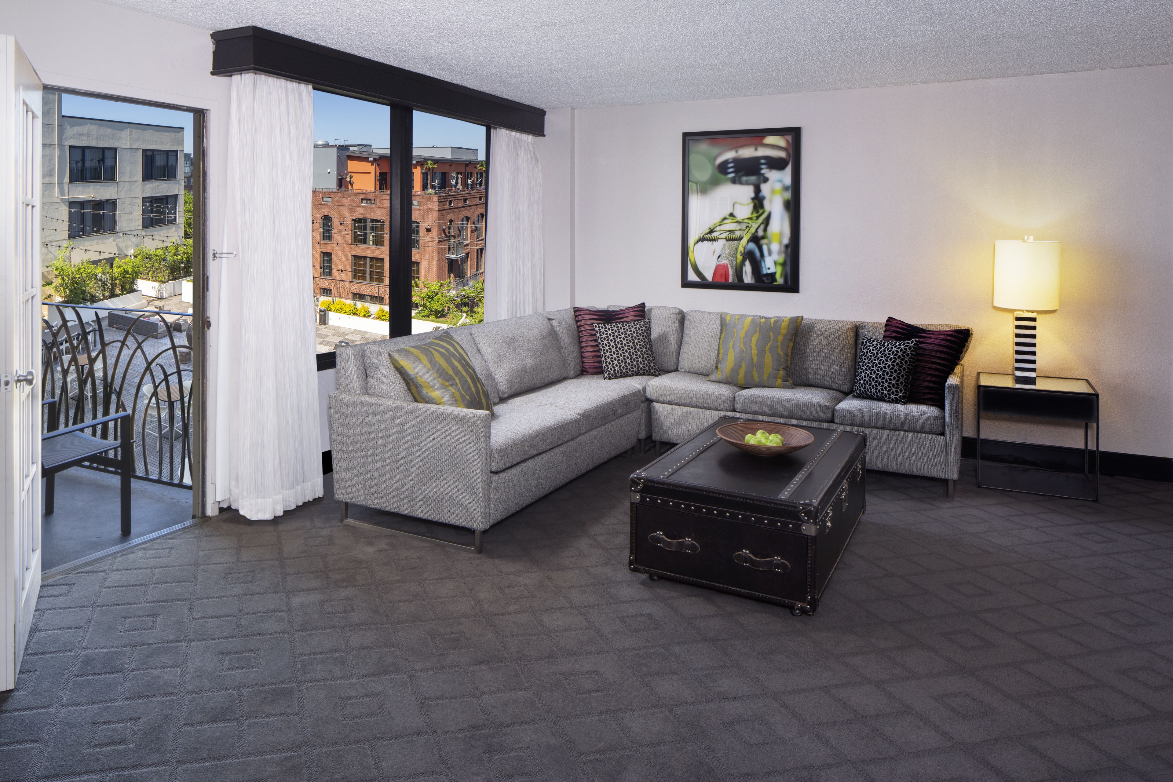 A modern living room with a grey sectional sofa, a black trunk, and a view of buildings through large windows.