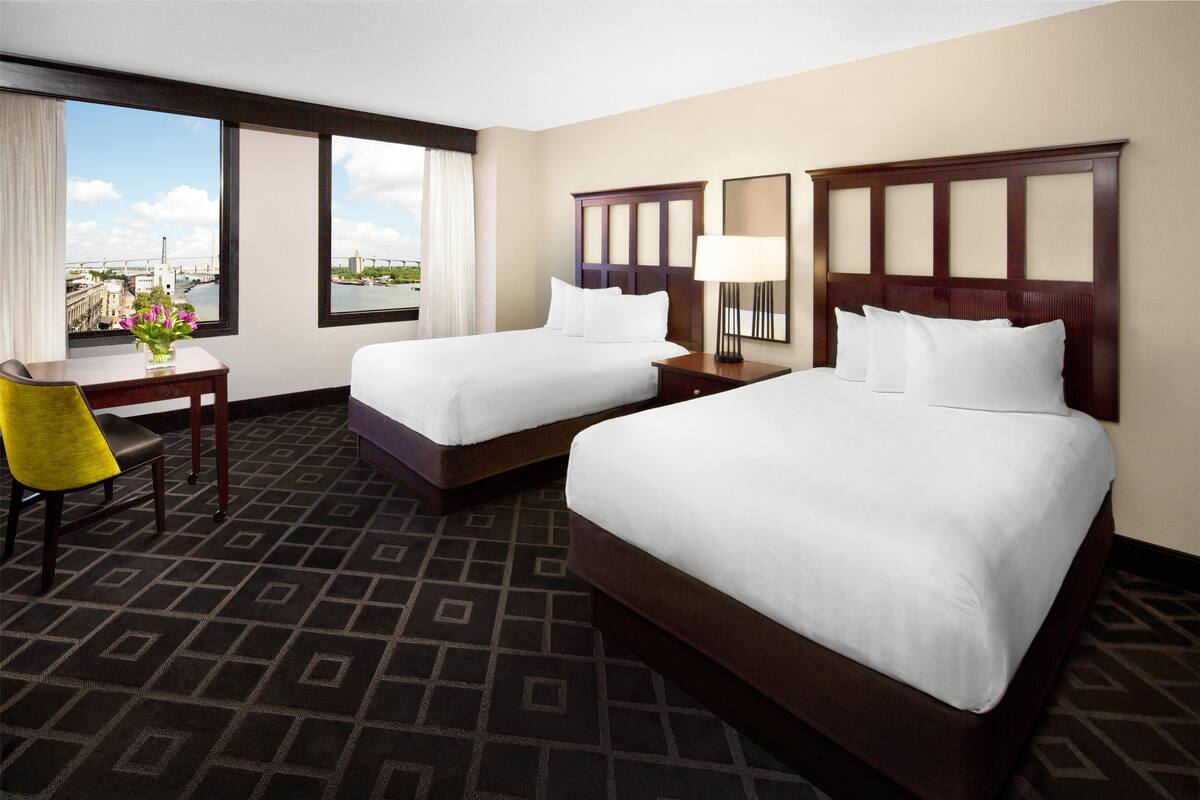 A hotel room with two beds, a desk, a chair, and a large window offering a city view.