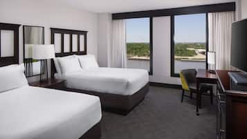 A hotel room with two beds, a desk, a chair, a TV, and large windows with a view of the outdoors.