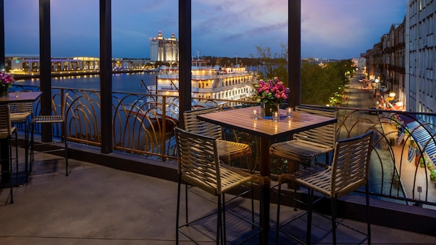 A balcony with wicker chairs and tables overlooking a river with boats and buildings in the background. - Hyatt Regency Savannah (Savannah)