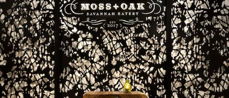 Una mesa de madera con una botella de vidrio, sobre un fondo abstracto en blanco y negro con el texto "Moss+Oak Savannah Eatery, Two West Bay St., Established 2017".