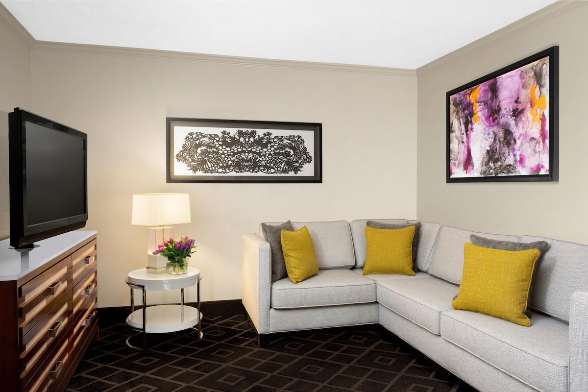 A modern living room with a grey sectional sofa, a flat-screen TV, a small side table with a lamp, and two framed artworks.