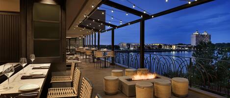 An outdoor dining area with tables and wicker chairs, a fire feature, and a view of a body of water and buildings at dusk.