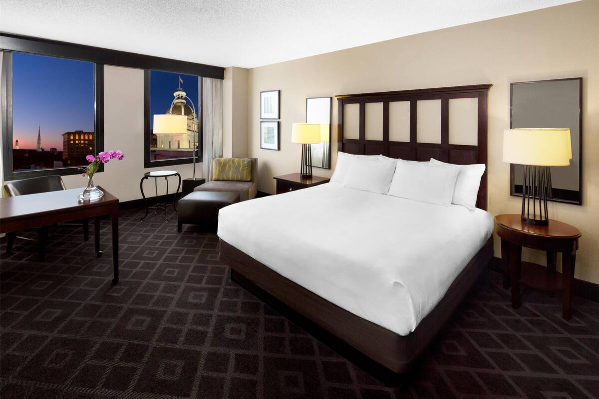 A hotel room with a large bed, a desk, a chair, a small table, and a window with a city view.