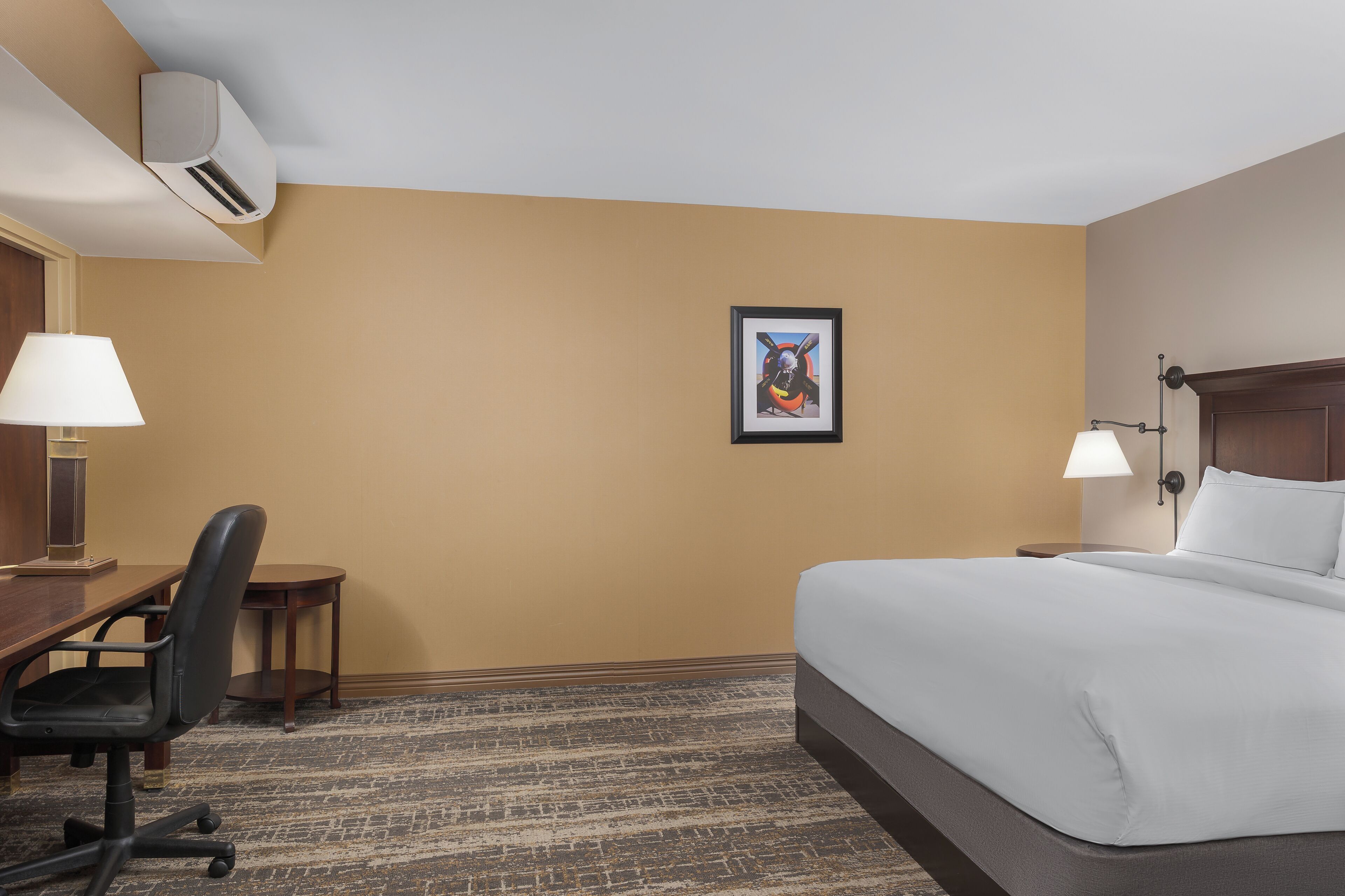 A hotel room with a bed, desk, chair, lamp, and framed artwork on the wall.