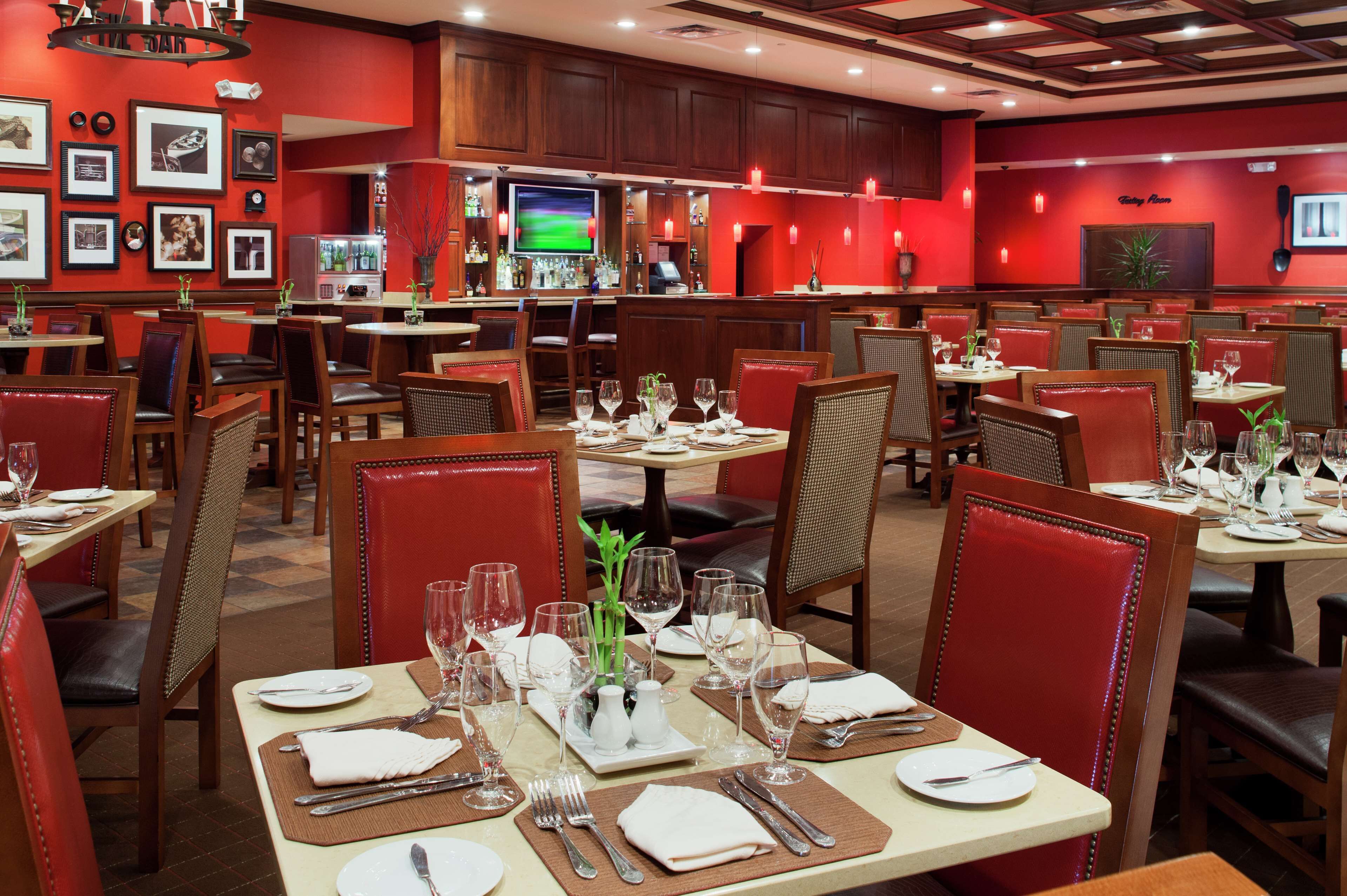 A restaurant with multiple dining tables set for guests, featuring red chairs and a bar area in the background.