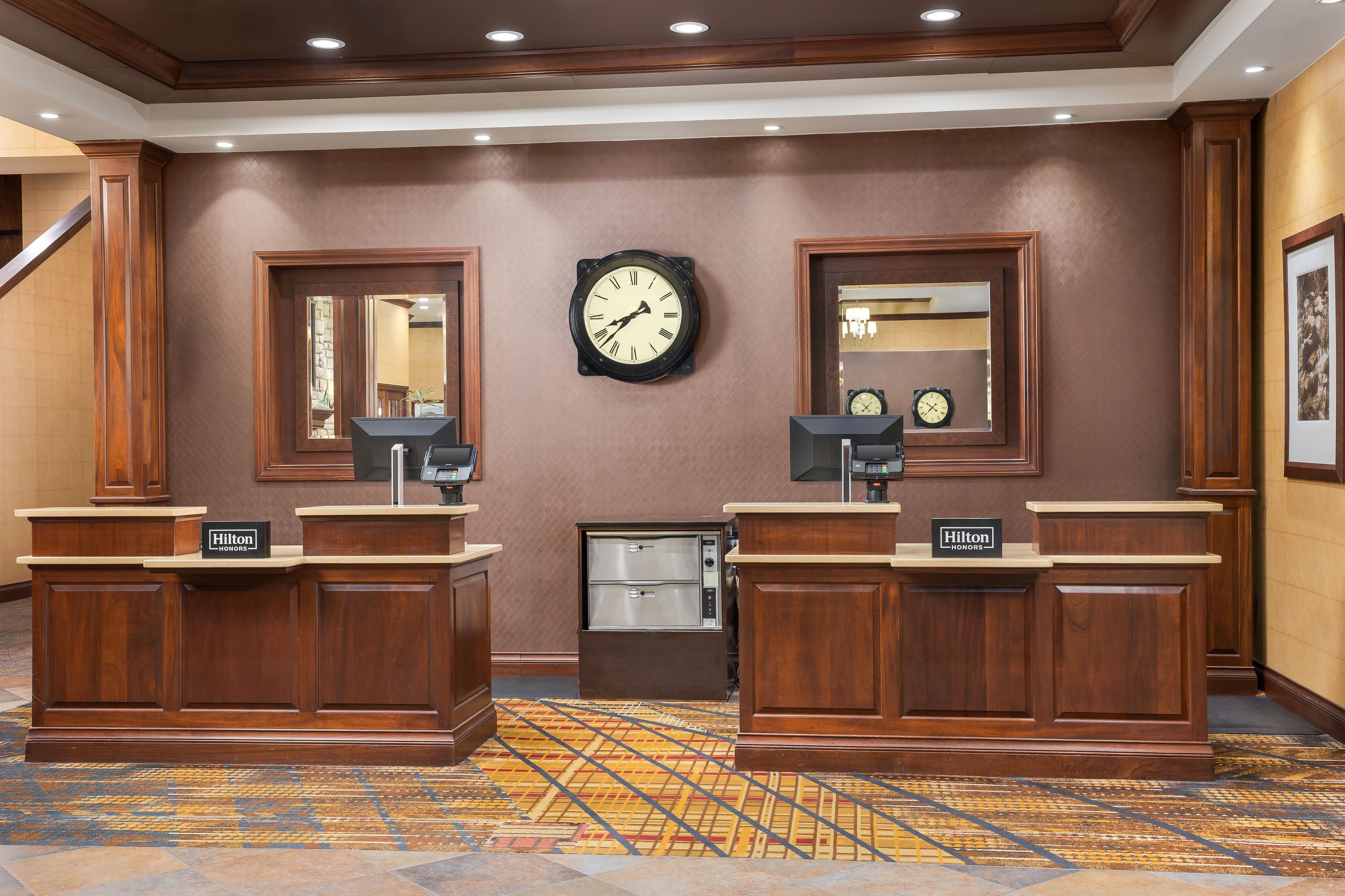 Un espace d’accueil d’hôtel comprenant deux comptoirs de réception en bois, une horloge murale et un miroir.