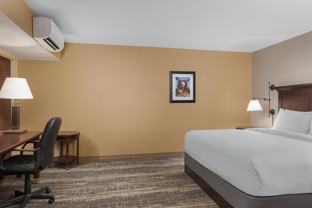 A hotel room with a bed, desk, chair, lamps, and an air conditioning unit.