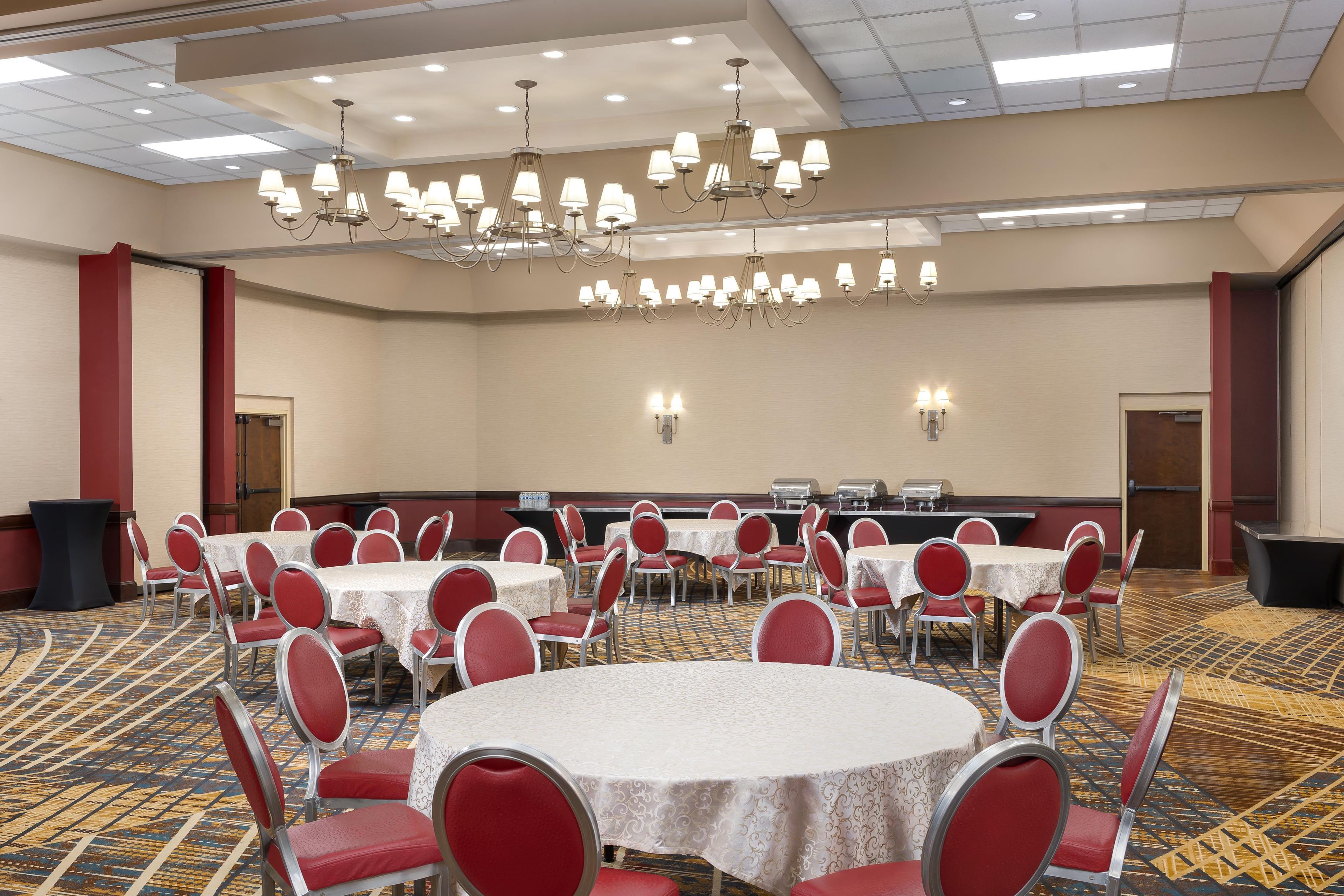 A conference room with multiple round tables, red chairs, a chandelier, and a carpeted floor.