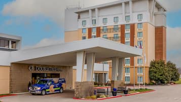 Un hotel con entrada techada, una camioneta estacionada al frente y un asta con banderas.