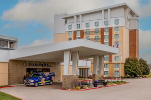 A hotel with a covered entrance, a van parked in front, and a flagpole with flags. - DoubleTree by Hilton Cincinnati Airport (Florence)