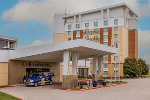 Un hotel con entrada techada, una camioneta estacionada al frente y un asta con banderas.