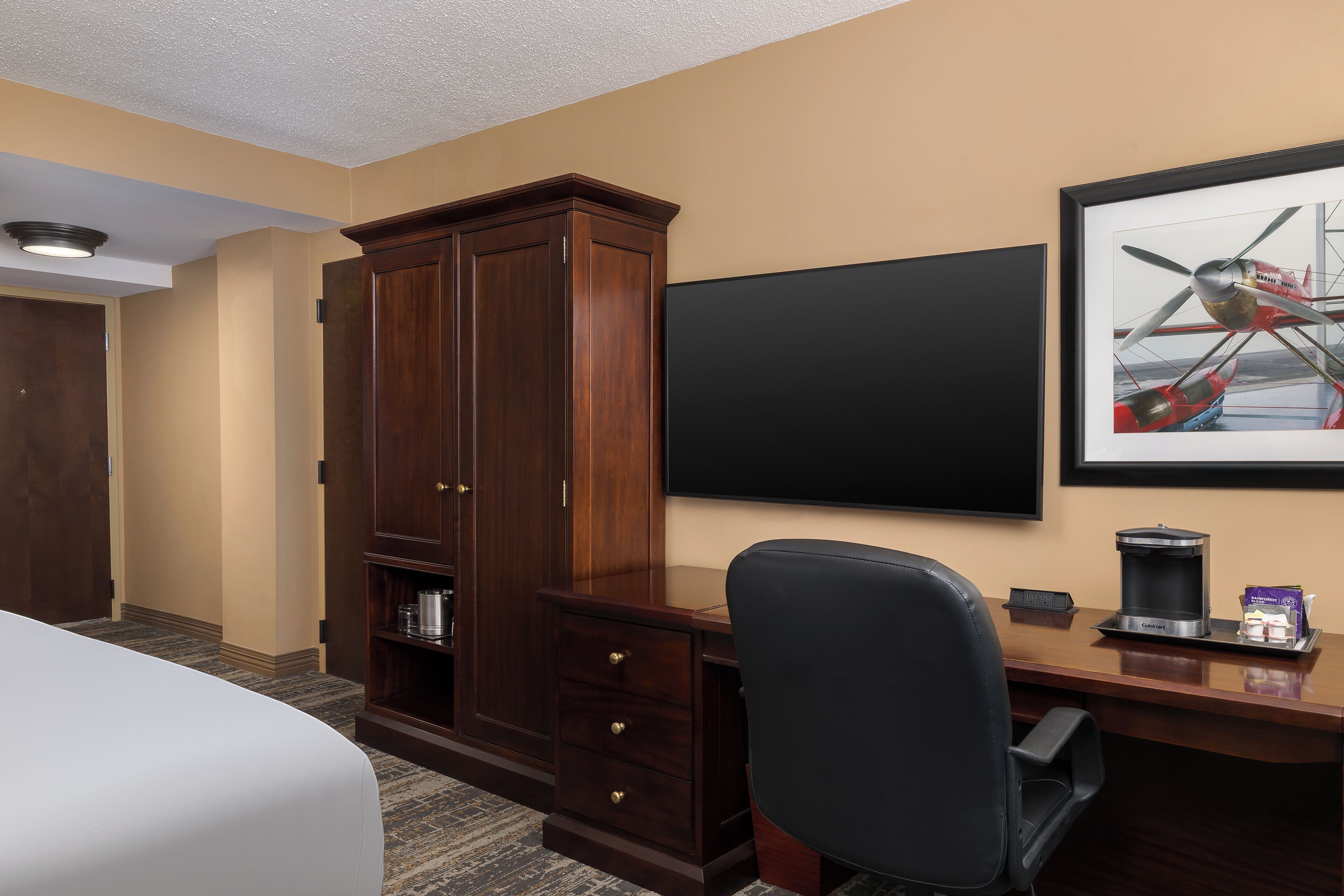 A hotel room with a large wooden cabinet, a desk with a black chair, a flat-screen TV, and a framed picture of an airplane.