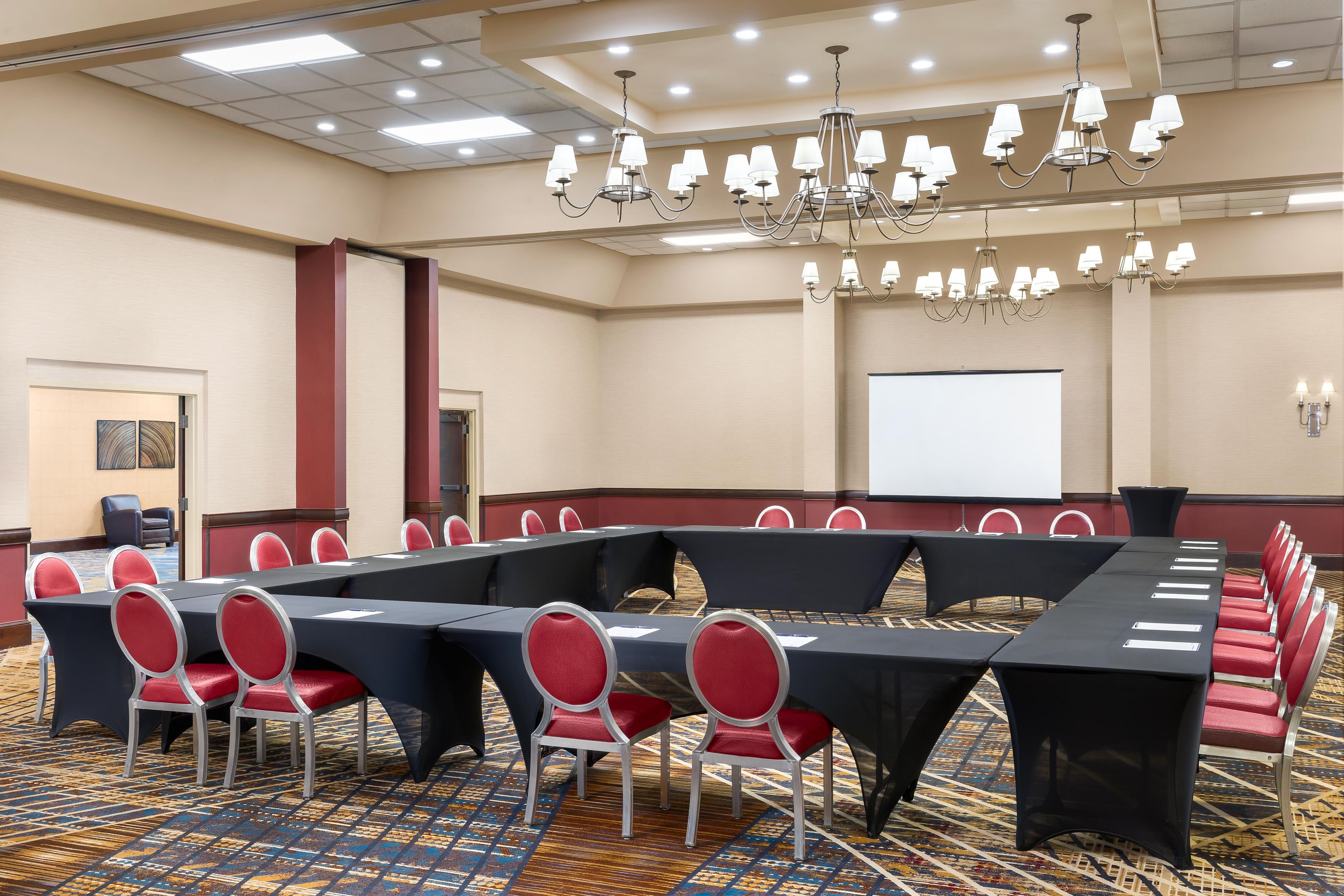 A conference room with a long table, red chairs, a projector screen, and multiple chandeliers.
