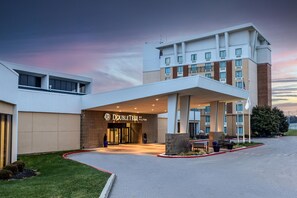 L’entrée d’un hôtel, avec un espace d’attente abrité, un ciel dégagé et une pelouse bien entretenue.