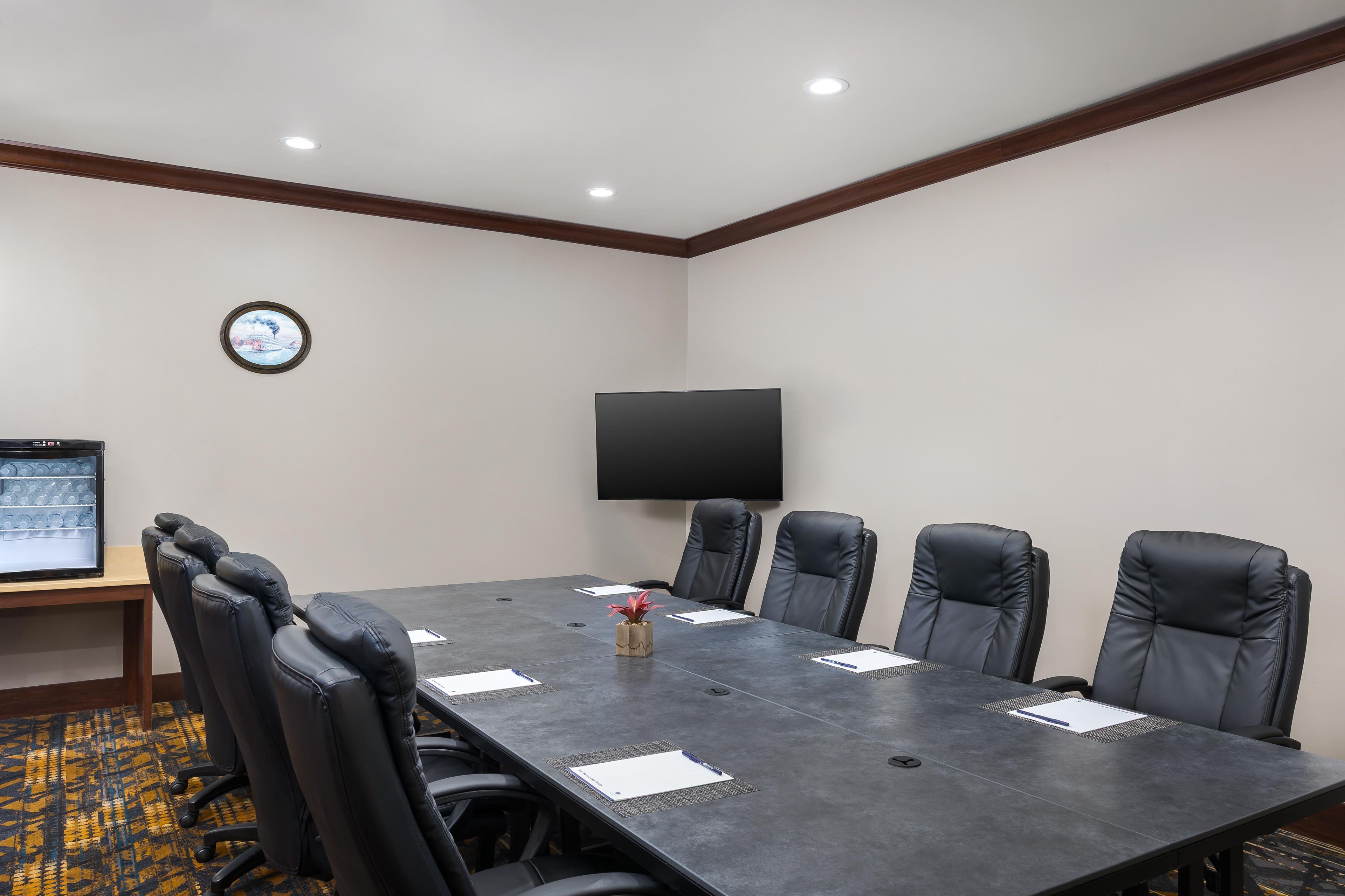 A conference room with a long table, six black leather chairs, a flat-screen TV mounted on the wall, and a small clock on the wall.