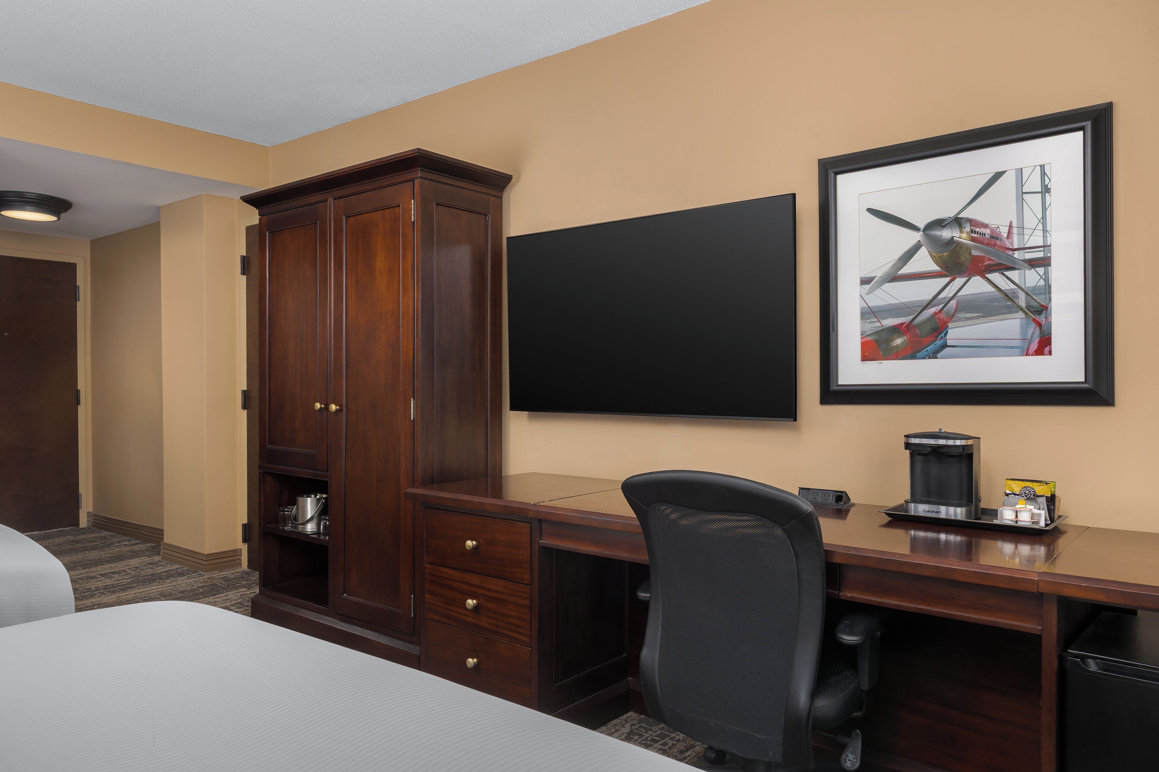 A hotel room with a wooden desk, a black chair, a flat-screen TV, a framed airplane picture, and a minibar.